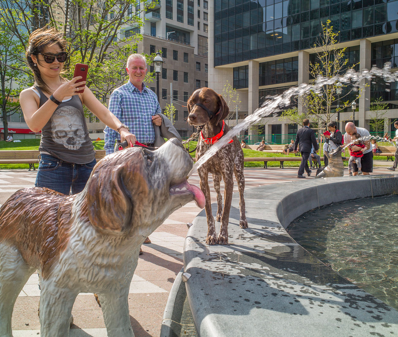 A young woman taking a picture of the fountain at Berczy Park in Toronto while her dog is looking at the statue